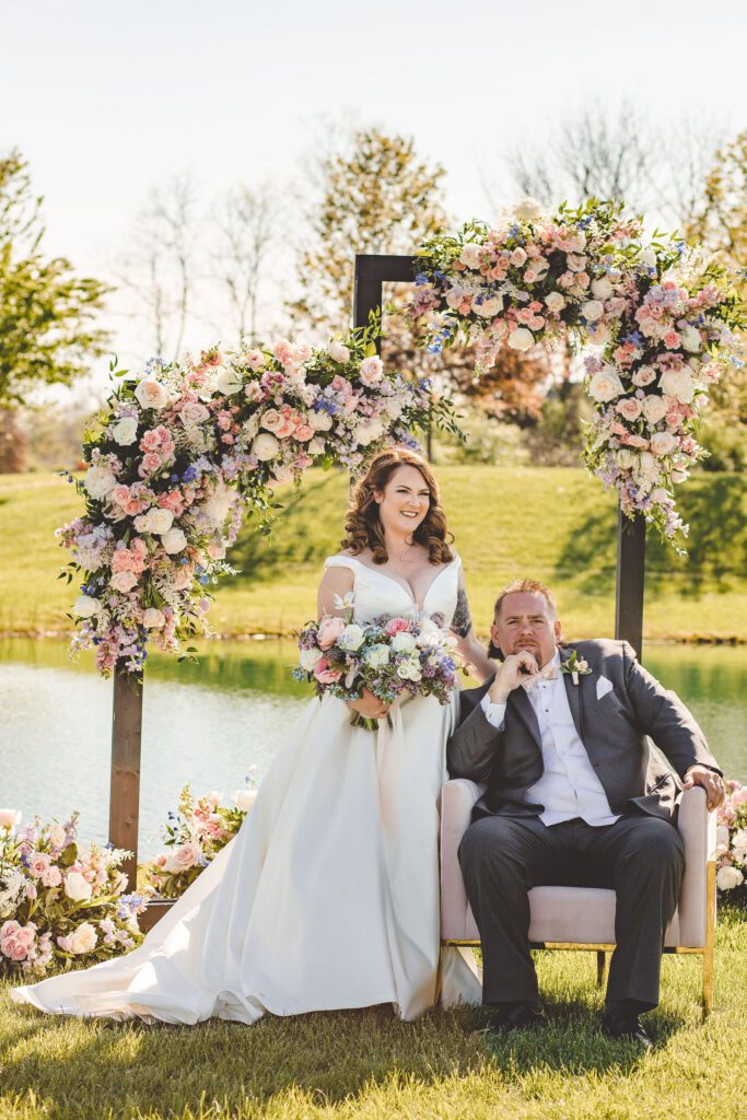Bride holding her bouquet next to her groom, who is seated in front of their flower covered wedding arches. 